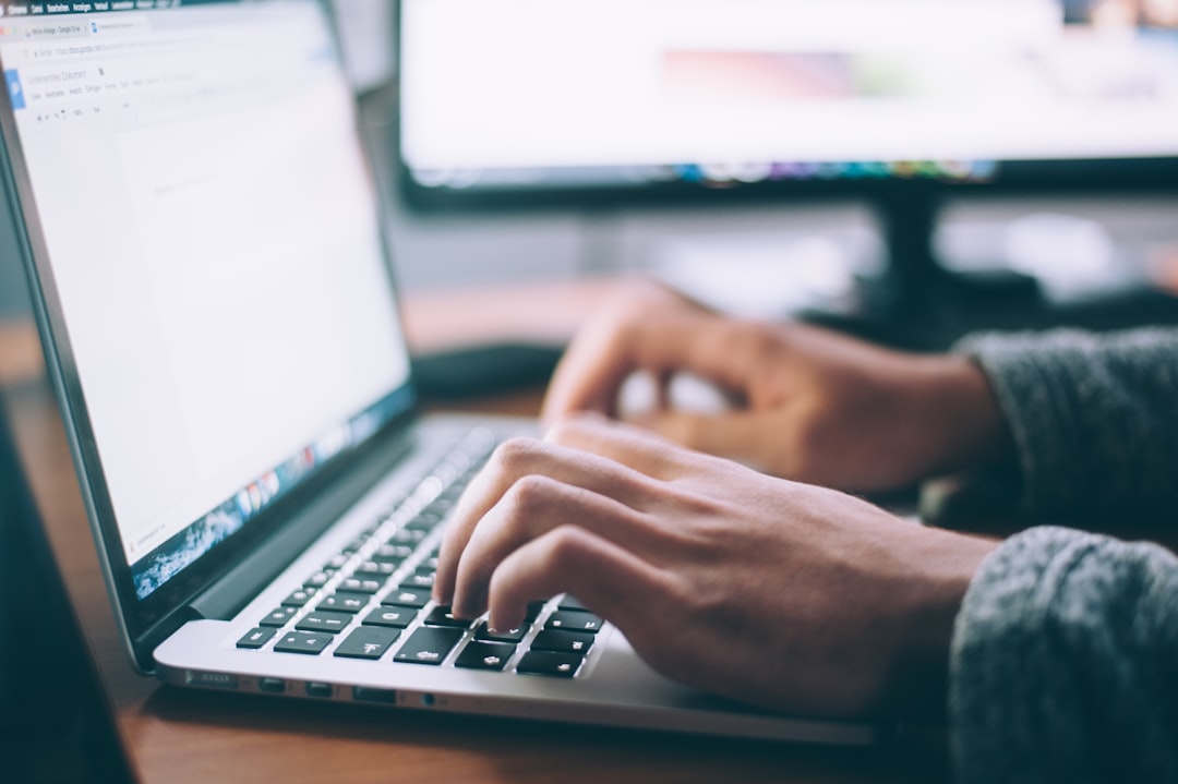 En person skriver på ett laptoptangentbord vid ett skrivbord, med en skärm och en suddig skärm i bakgrunden, vilket antyder en arbets- eller studiemiljö fokuserad på WordPress-hostinguppgifter.